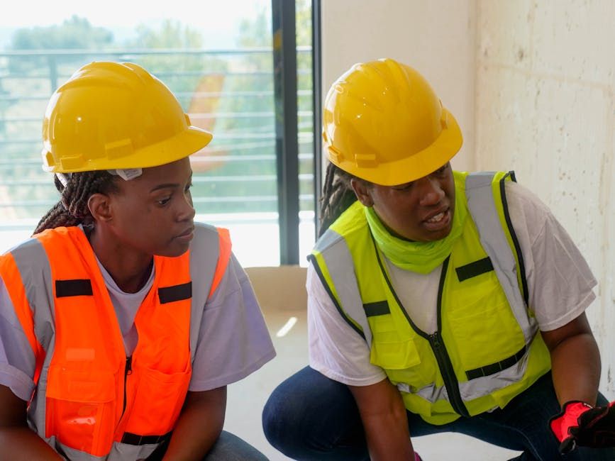 Two engineers wearing protective gear and hardhats discuss a project inside a building under construction.